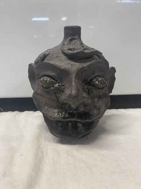 A dark, textured ceramic Face Jug Attributed to DX Gordy with exaggerated facial features displayed on a white cloth background.
