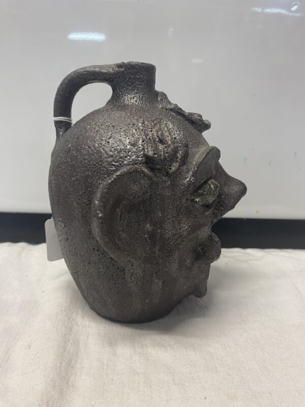 A dark ceramic Face Jug attributed to DX Gordy, shaped like a human head with pronounced facial features, displayed on a white surface.
