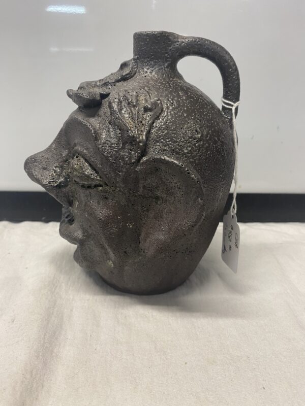 The Face Jug Attributed to DX Gordy is a textured ceramic jug with a whimsical human face design and a handle, complete with an attached price tag.