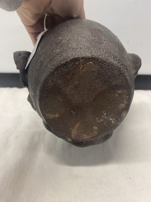 A hand holds the Face Jug Attributed to DX Gordy, an old, textured vessel with a round bottom, seen from its base.